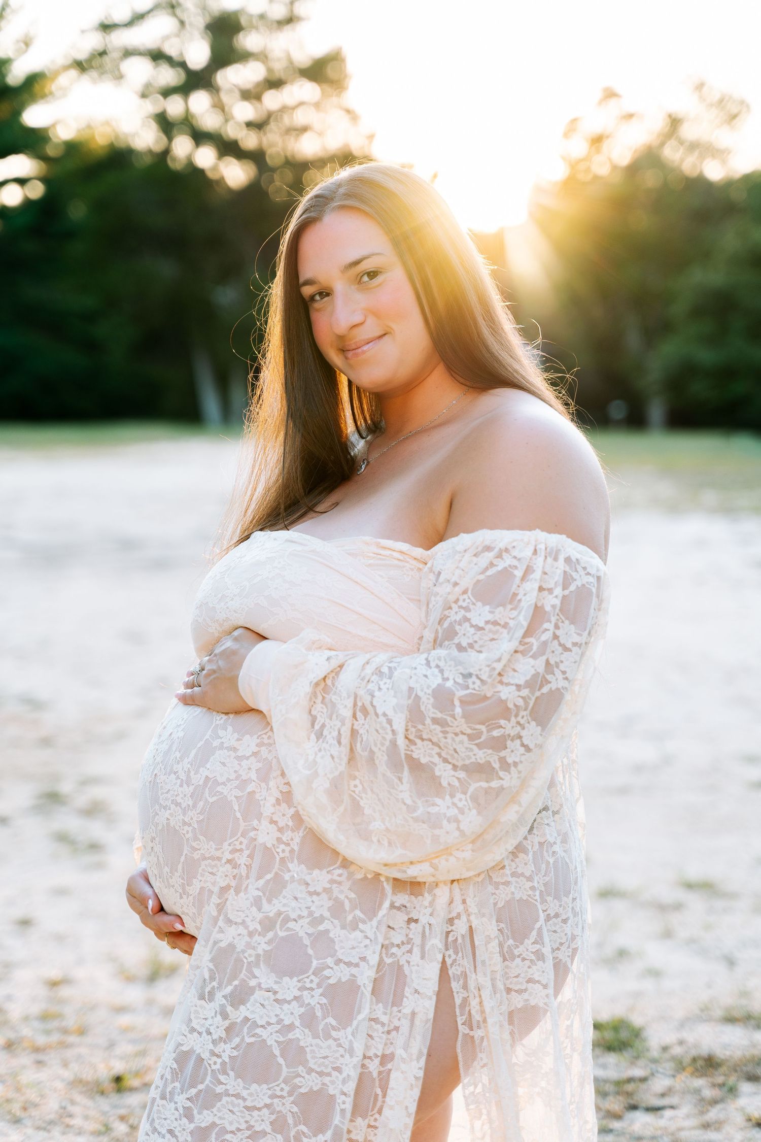 Pregnant woman in white lace off-shoulder gown poses outdoors at golden hour sunset with trees in background.