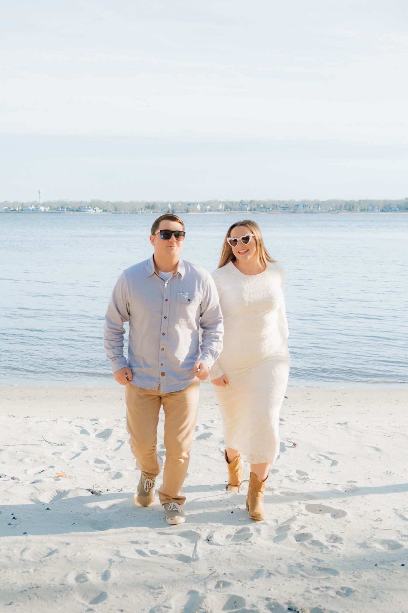 Couple walking hand in hand on a sandy beach, wearing sunglasses and casual outfits, with calm blue water in the background.