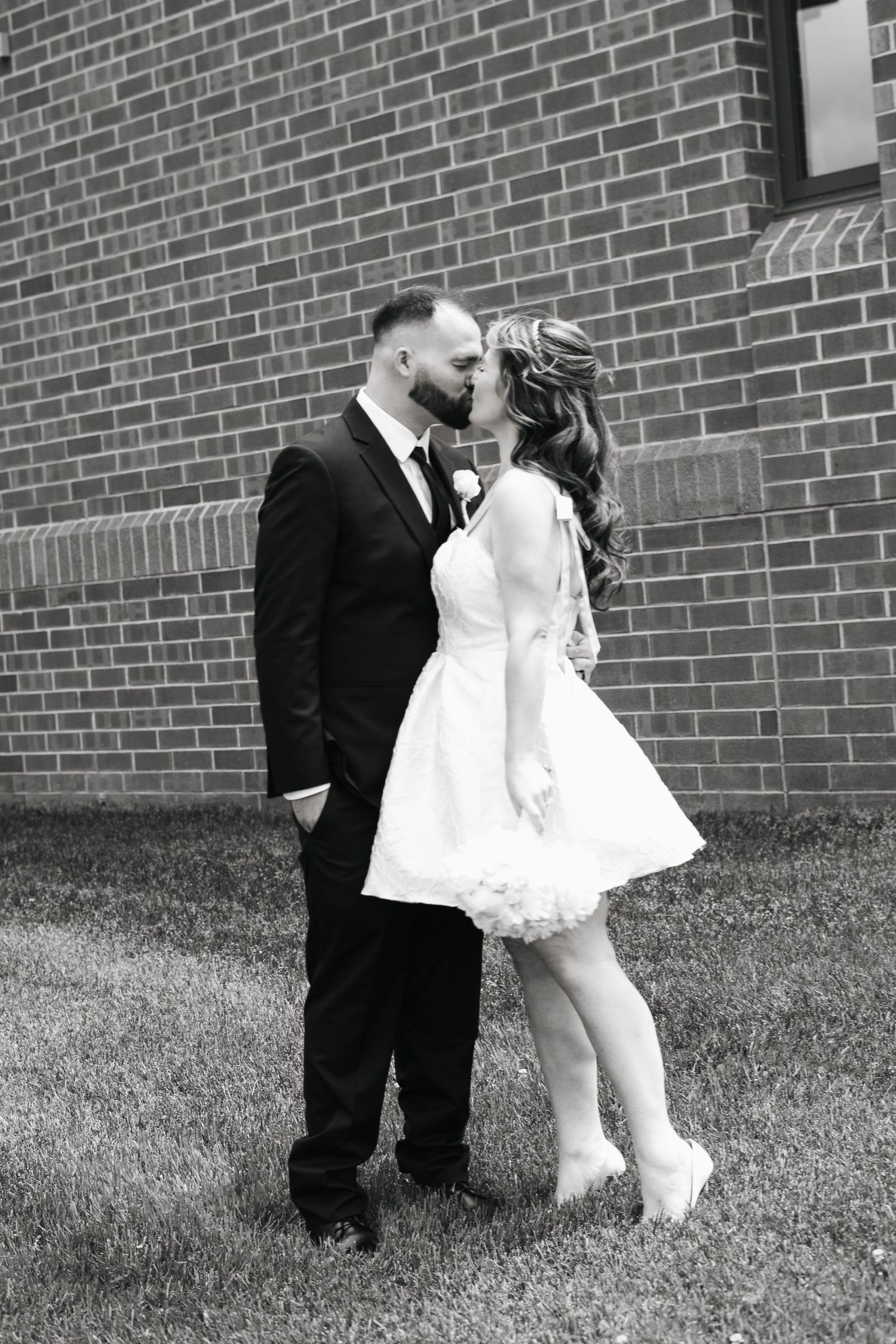 Black and white photo of bride in short white dress and groom in black tux sharing a kiss outside brick building.
