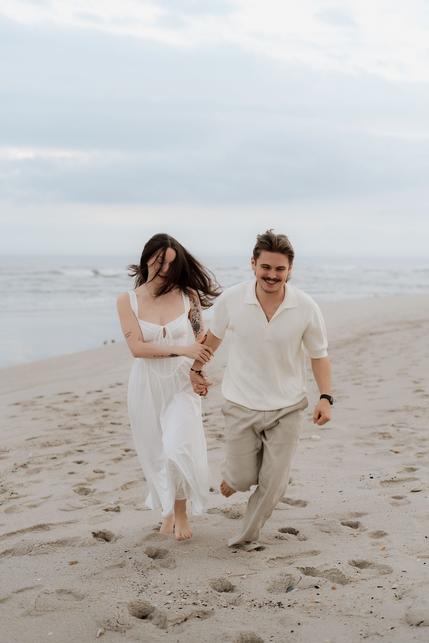 Happy couple running and laughing on sandy beach, woman in white dress, man in white shirt and tan pants holding hands.