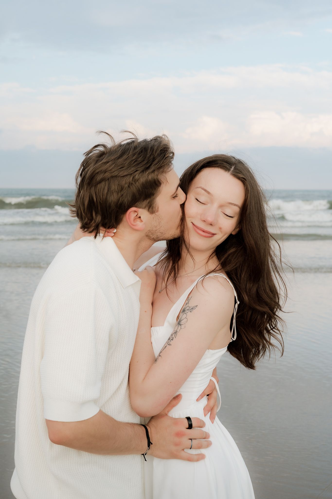 A couple embraces on the beach at sunset, the man kissing the woman's cheek as she smiles with eyes closed.
