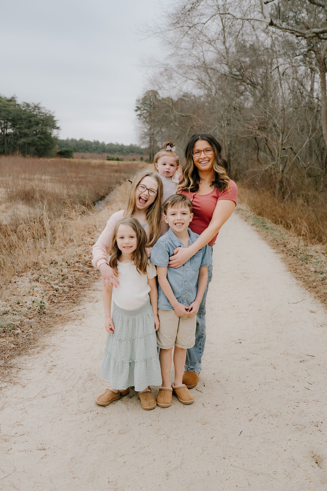 A laughing mother poses with her four children on a sandy woodland trail in casual outfits.