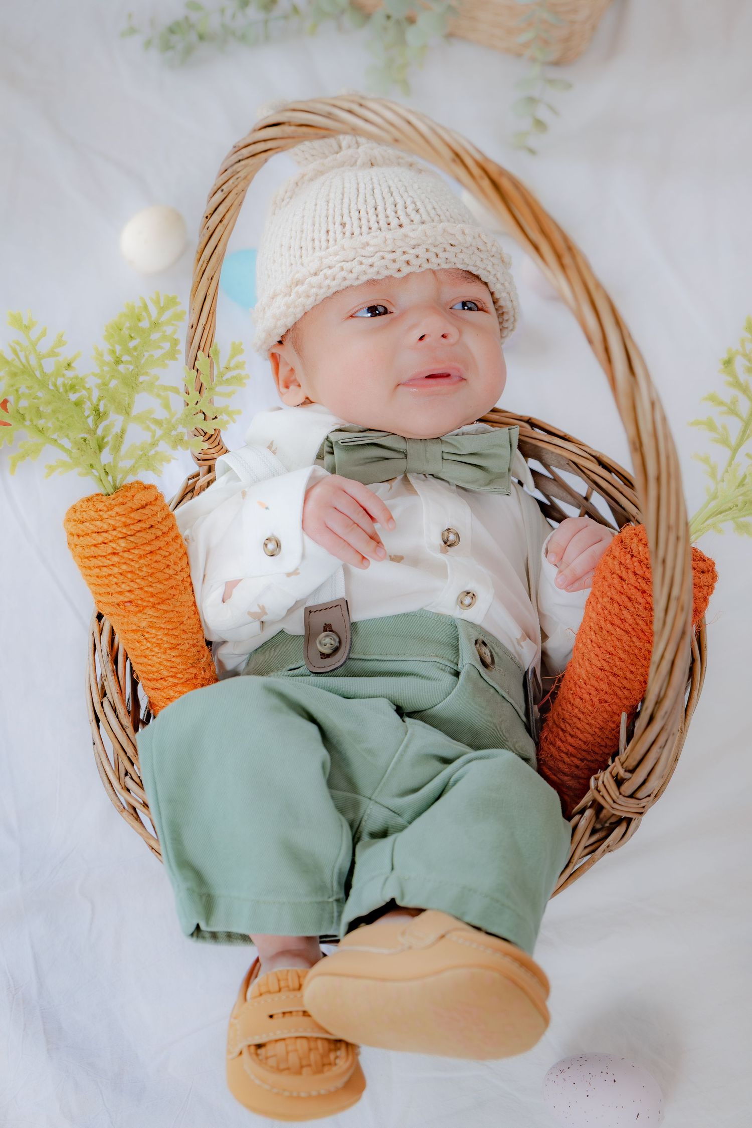 A newborn in a green outfit and bunny hat rests in a wicker basket with toy carrots.