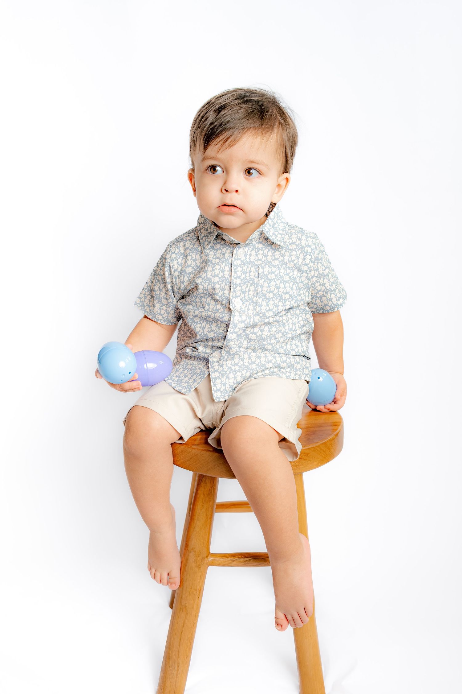 A cute toddler boy in a floral shirt and khaki shorts sits on a wooden stool holding colorful Easter eggs.