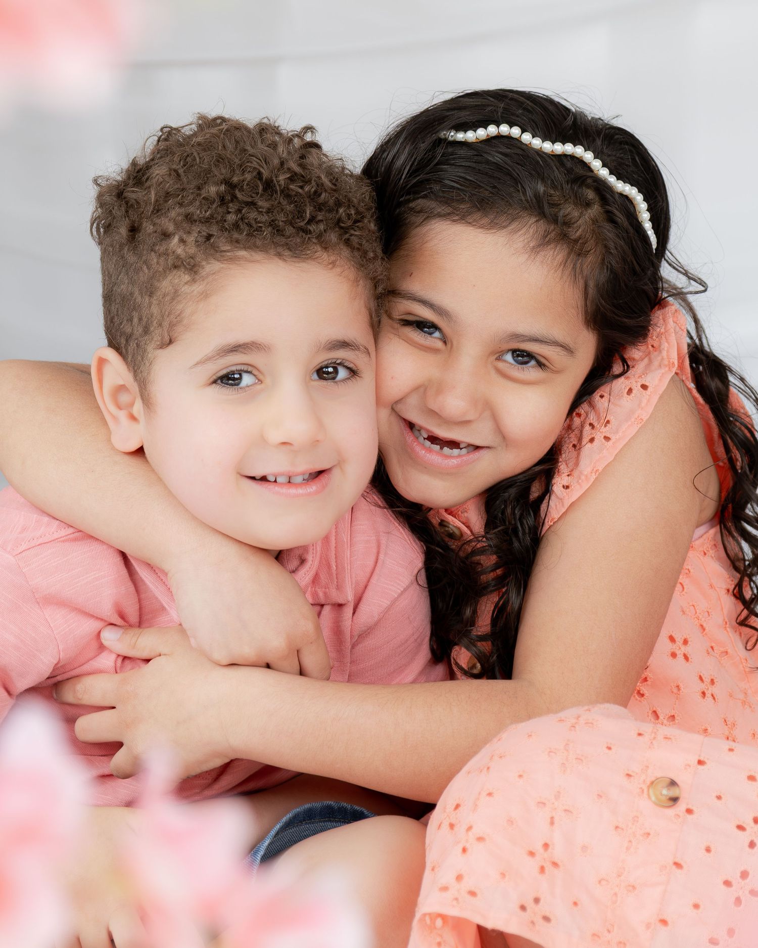 Two young siblings in coral outfits share a warm hug in a bright studio portrait.