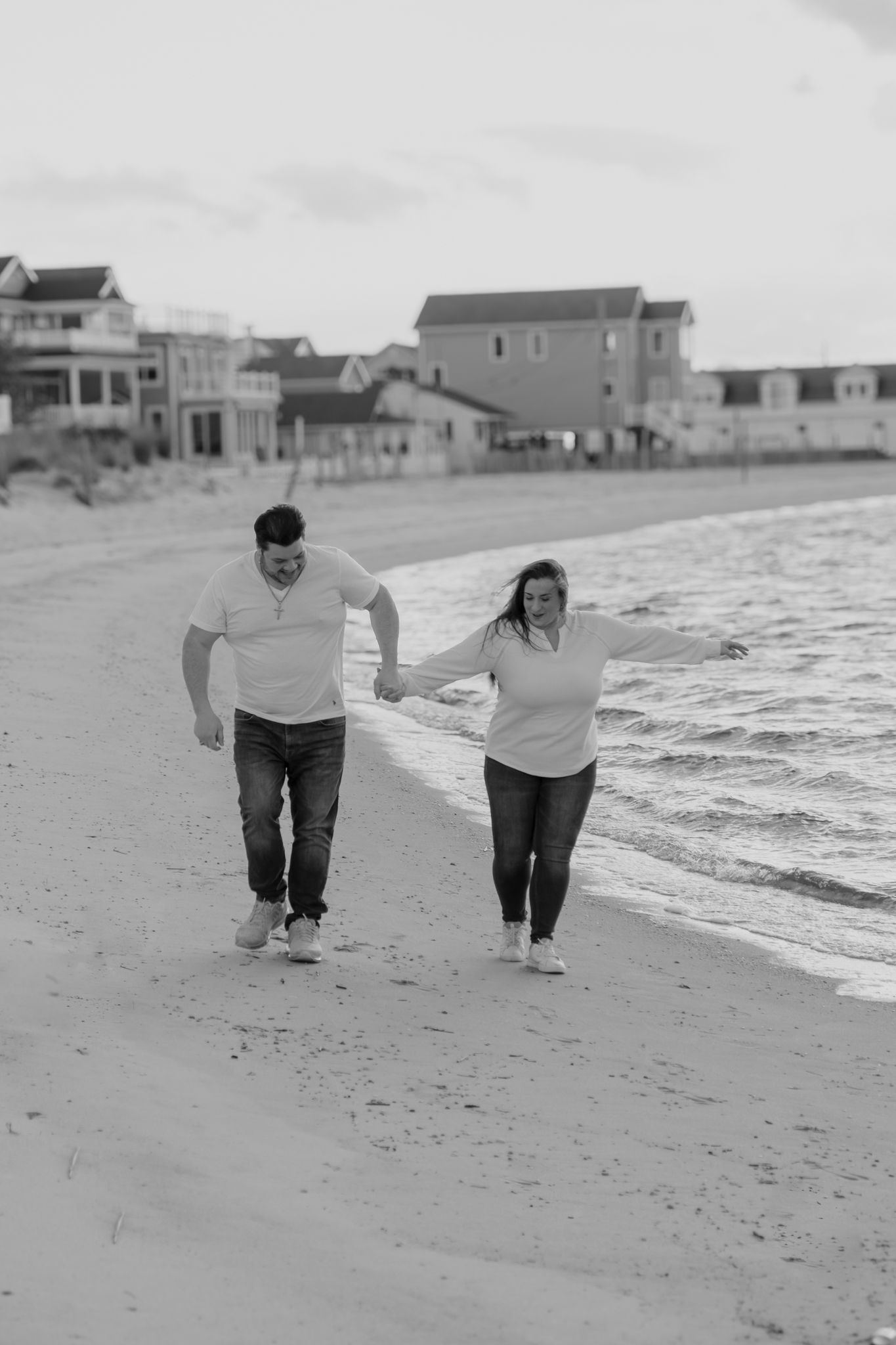 Black and white photo of a couple holding hands and walking along a sandy beach with coastal homes in the background.