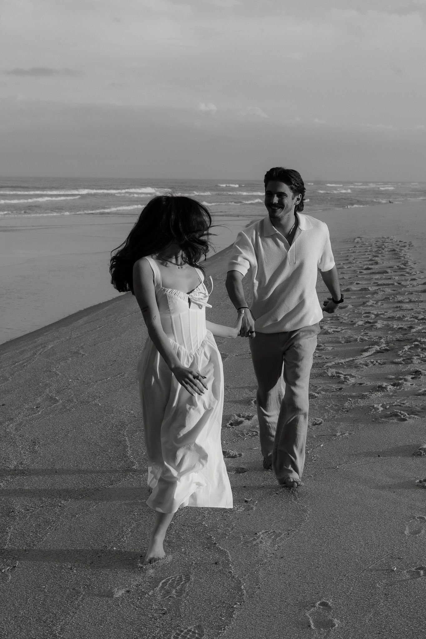 Black and white photo of a couple holding hands and walking barefoot on a sandy beach near ocean waves.