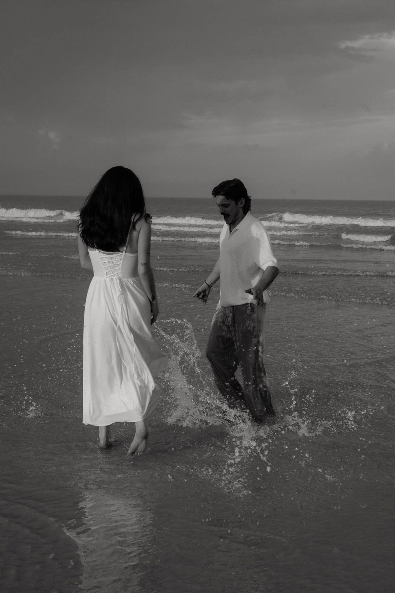 Black and white photo of a couple playfully splashing in ocean waves on a beach, woman in white dress.