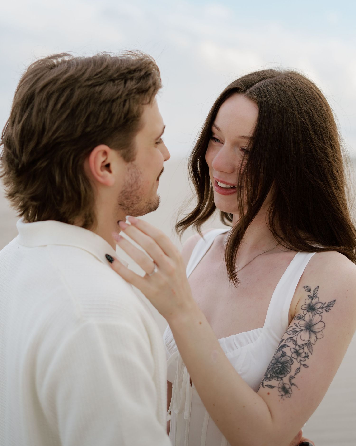 A couple shares a romantic moment, the woman smiling with a floral arm tattoo, both dressed in white outdoors.