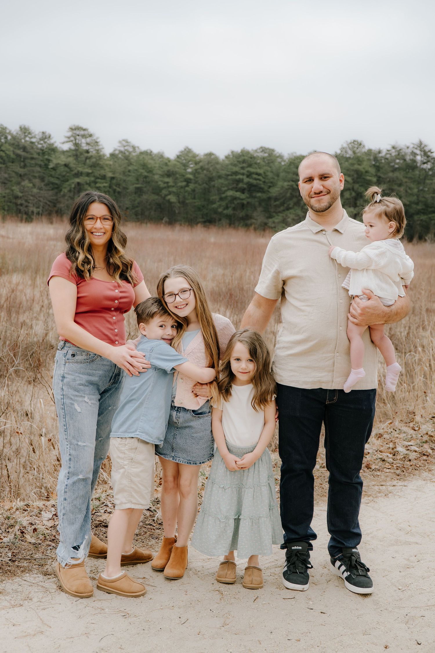 Family of six posing on a dirt path surrounded by tall grass and pine trees during an outdoor photo session.