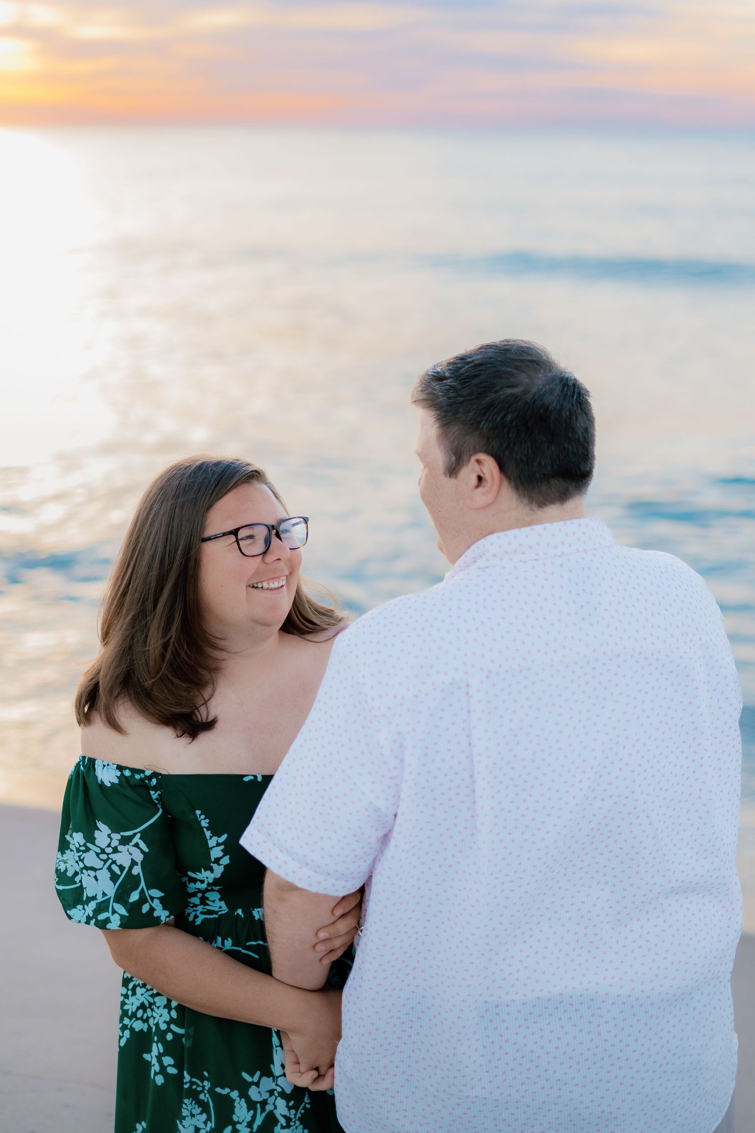 Couple holding hands on beach at sunset, woman in green floral dress with glasses smiling at man in white shirt.