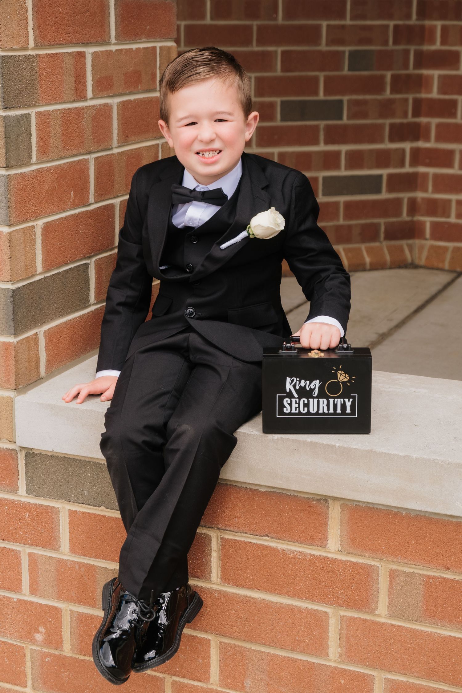 Smiling boy in black tuxedo holding a Ring Security briefcase with a white boutonniere at a wedding.