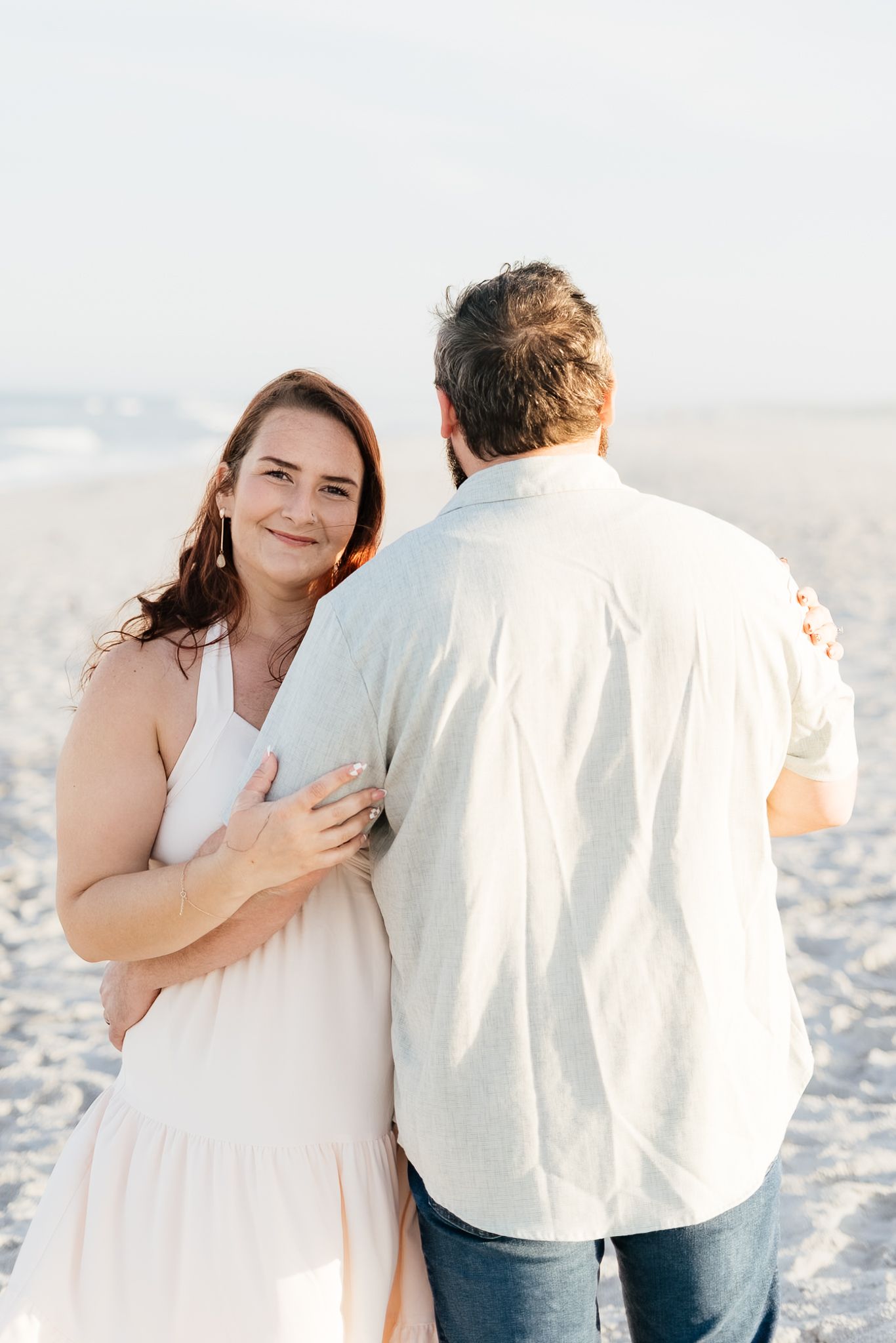 A couple embraces on a sandy beach, the woman in a white dress smiling warmly over her shoulder.