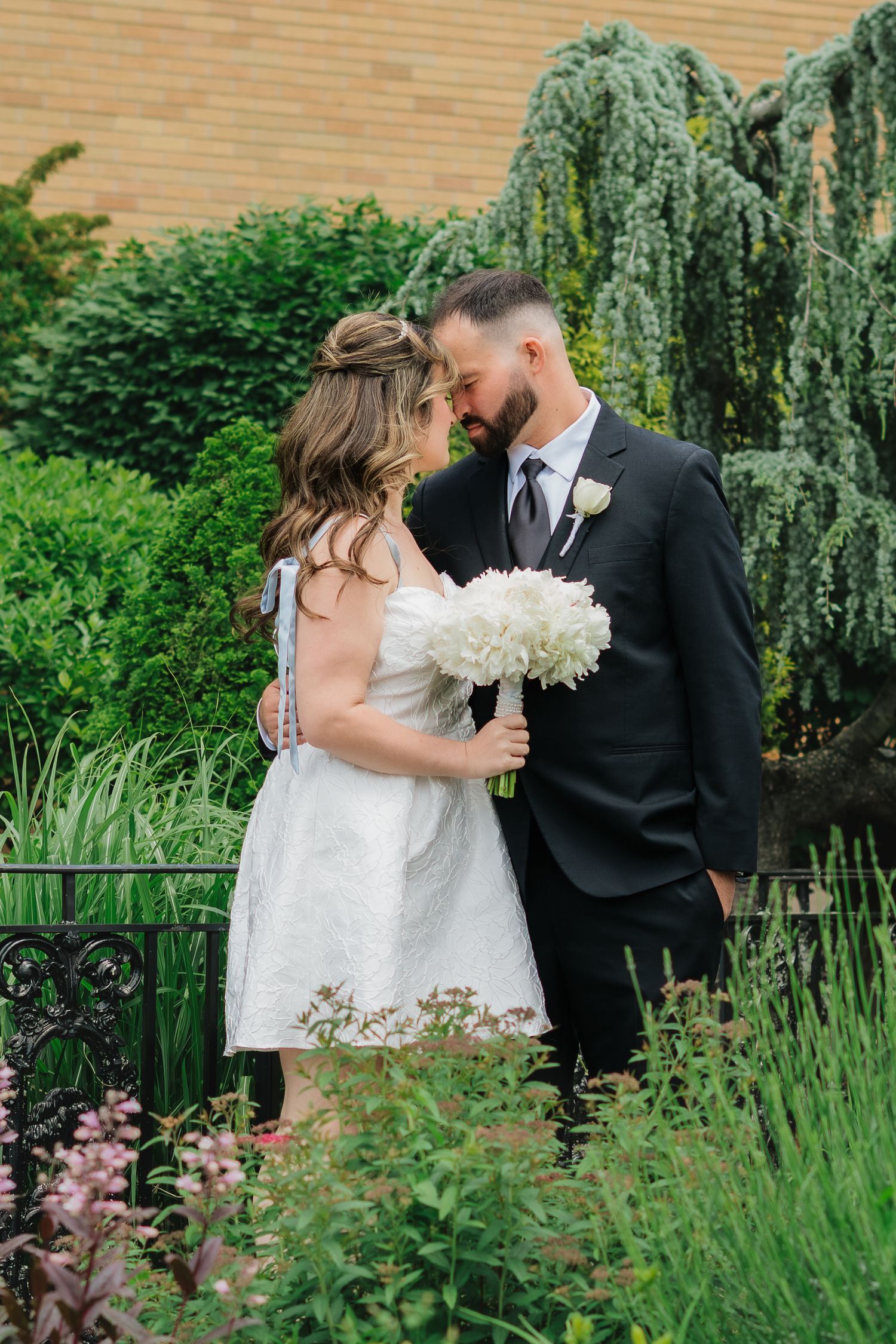 Bride and groom share a romantic moment in a lush garden, bride holding white bouquet in short wedding dress.
