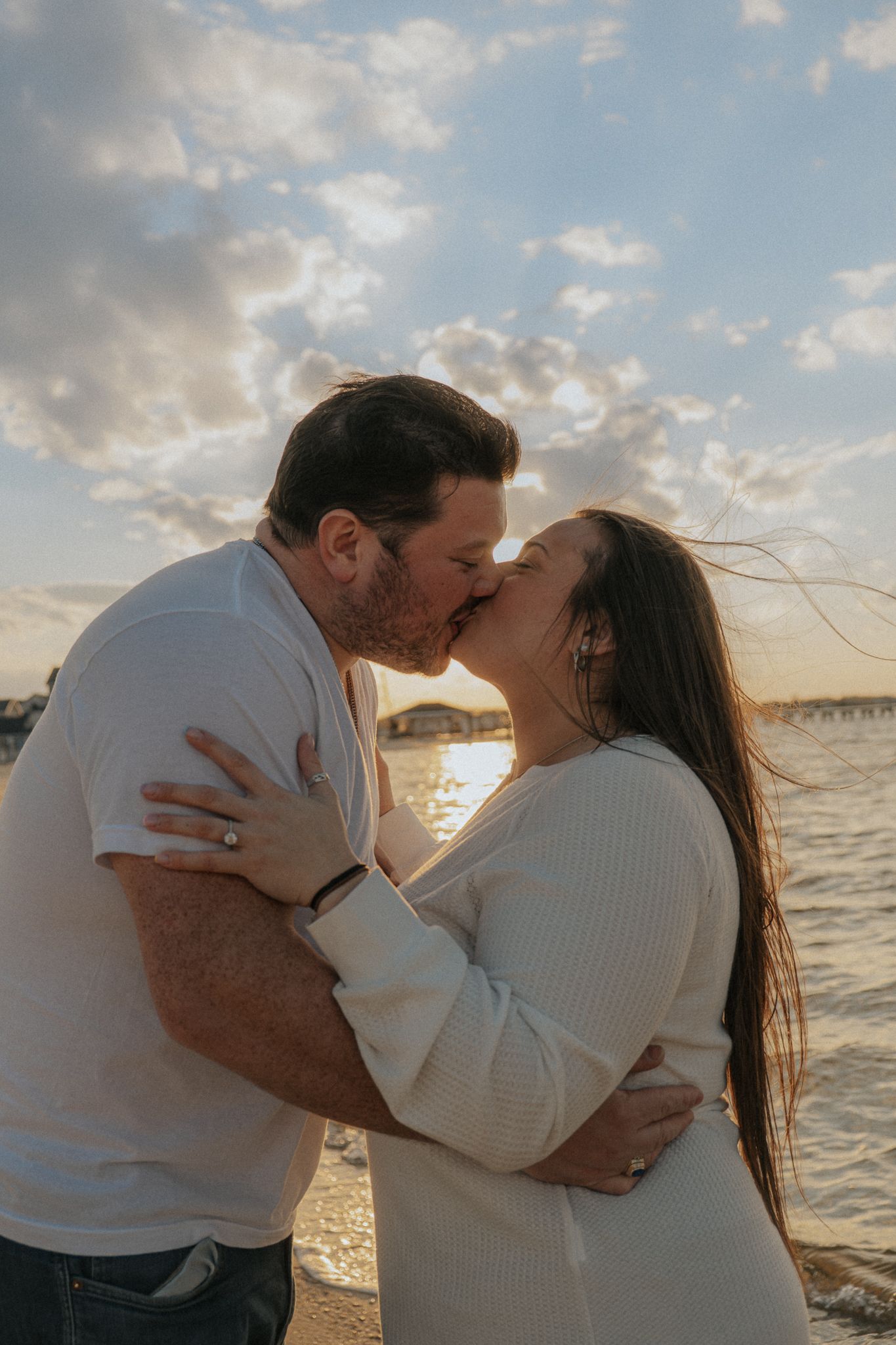Couple sharing a romantic kiss at sunset on the beach, embracing each other with golden light reflecting off the water.