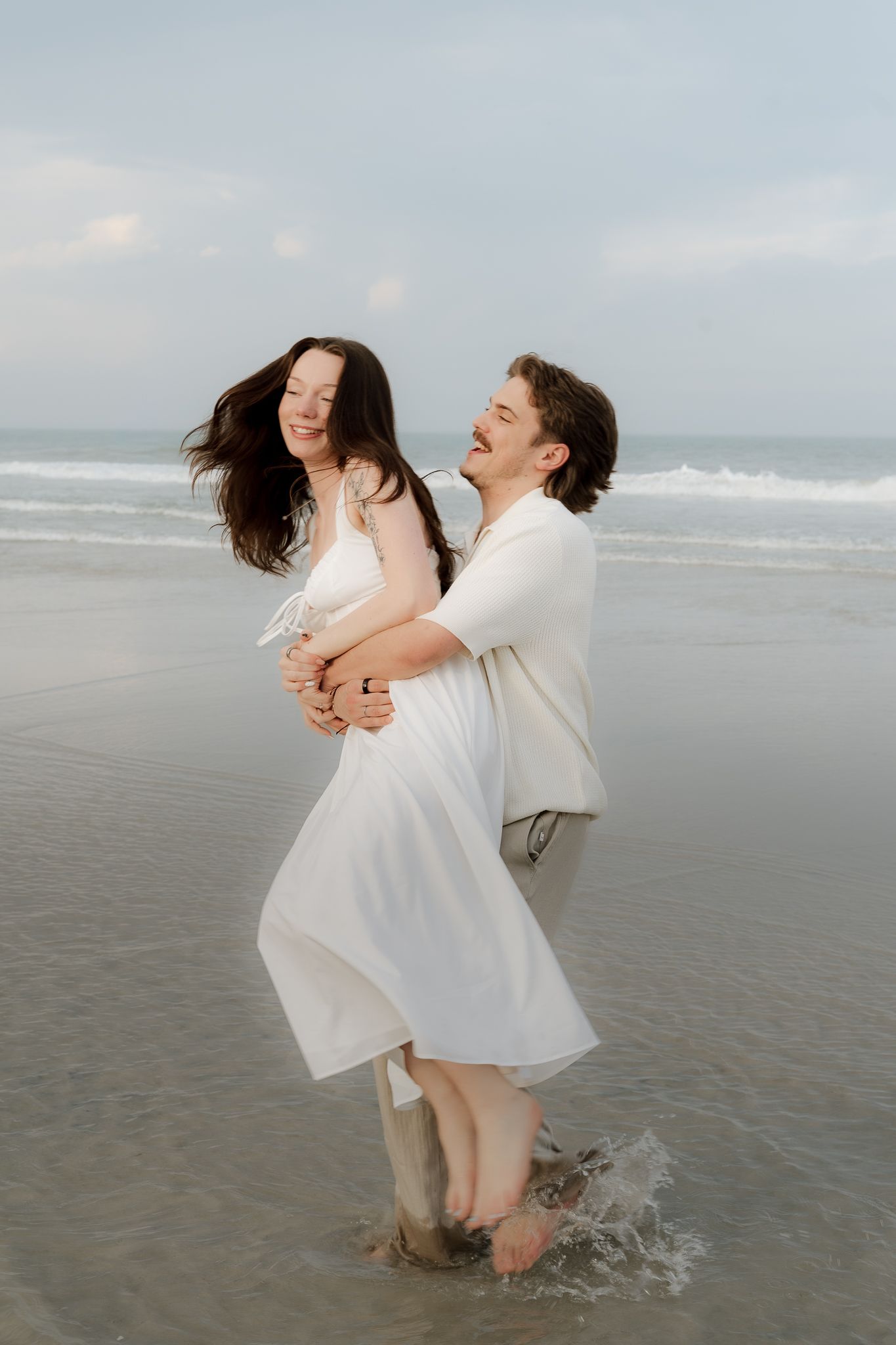 Joyful couple embracing and laughing in shallow ocean waves, woman in white dress lifted by smiling man.