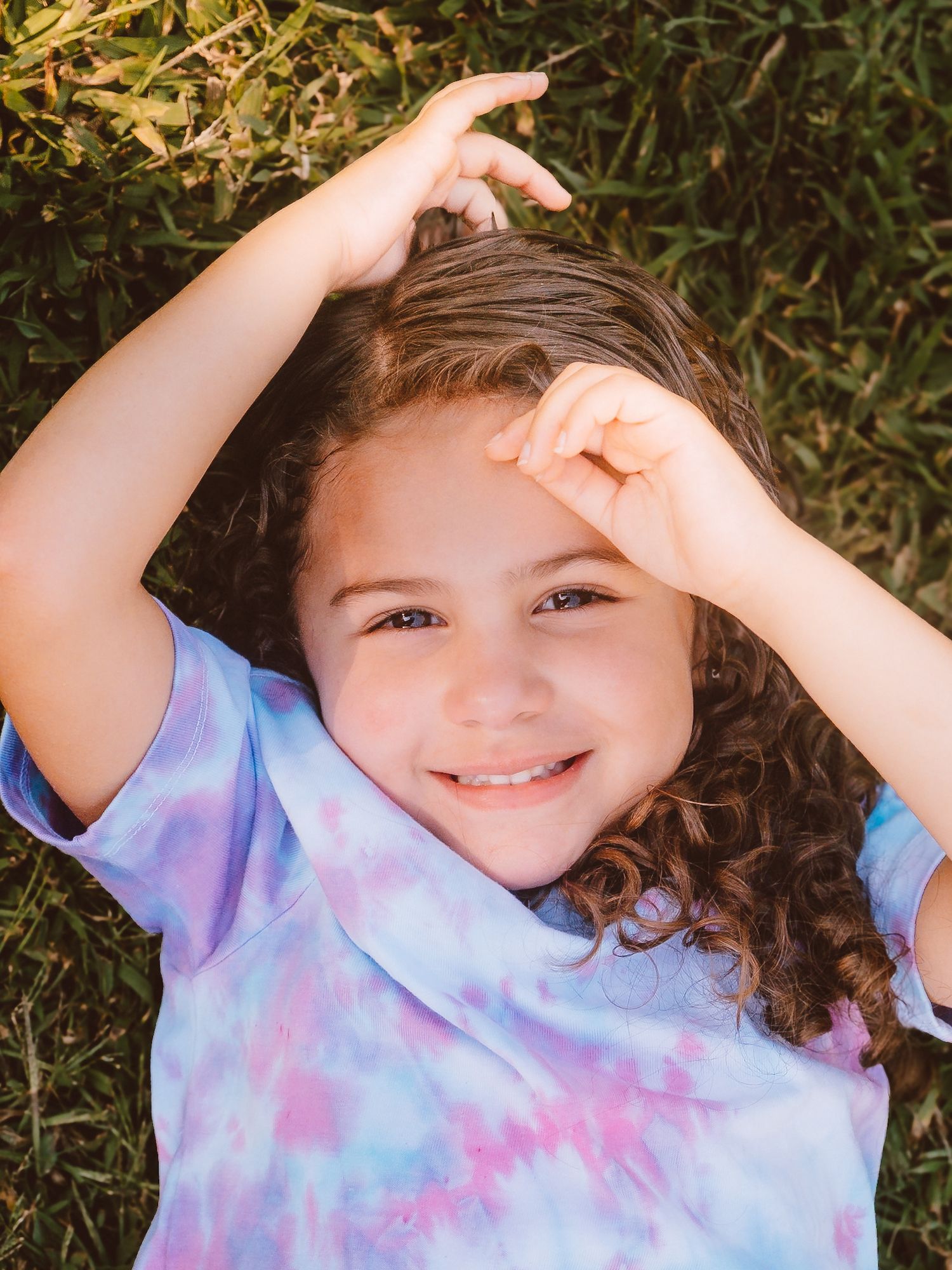 Smiling curly-haired girl in tie-dye shirt lying on grass with hands raised near her head.
