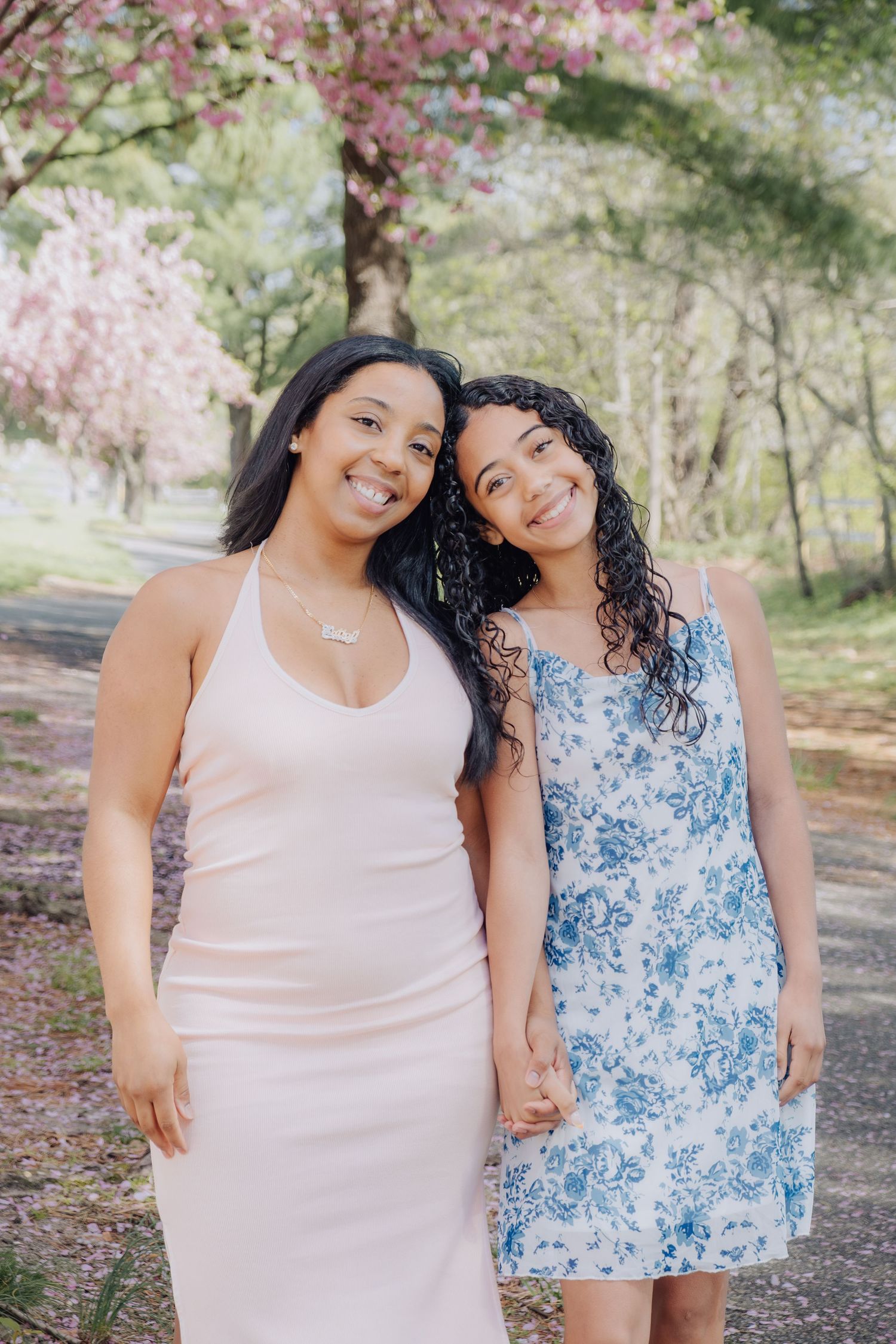 Two young women in spring dresses smile together outdoors near blooming pink cherry blossom trees.