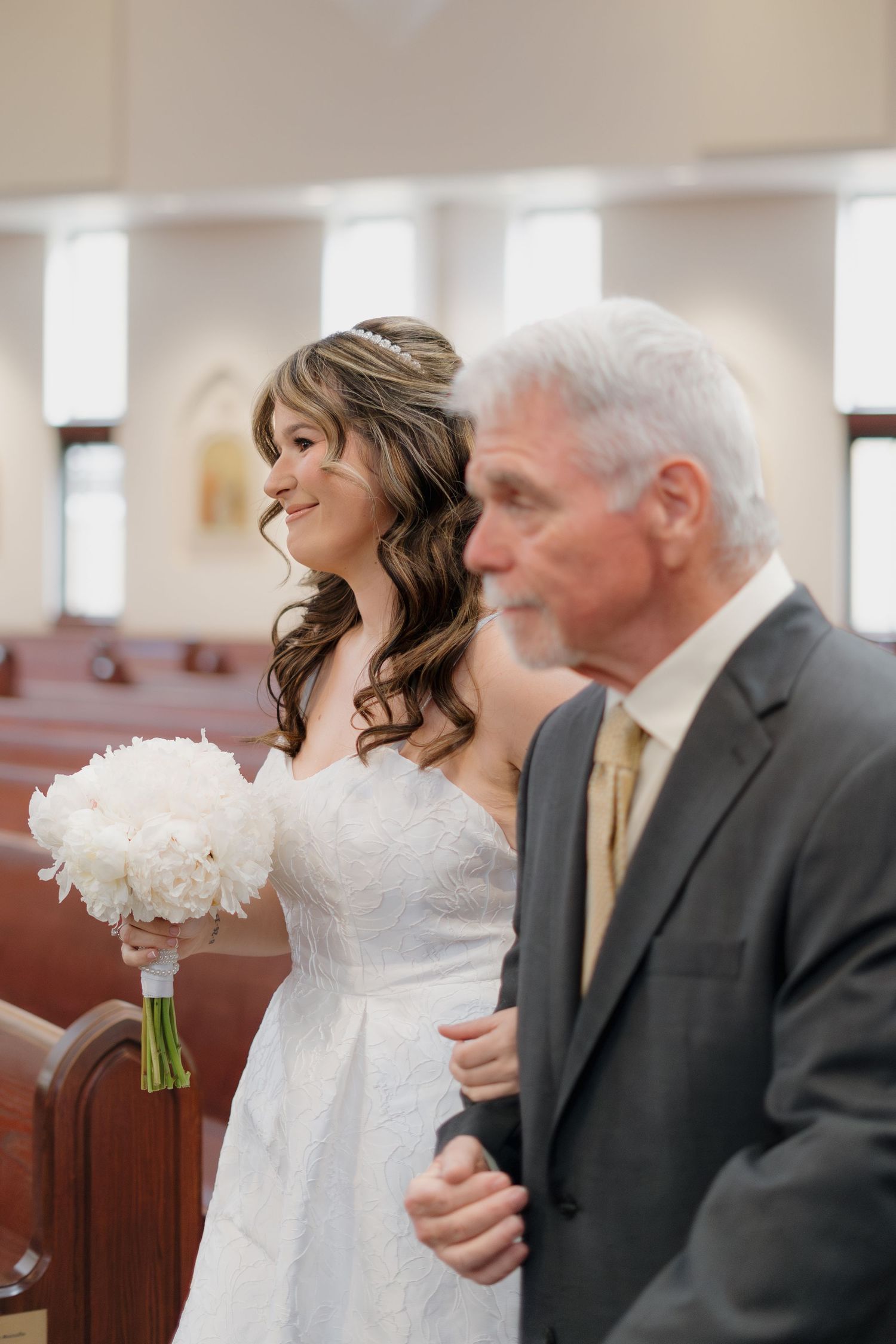 Radiant bride in white lace gown holding white peony bouquet walks down church aisle with her father.