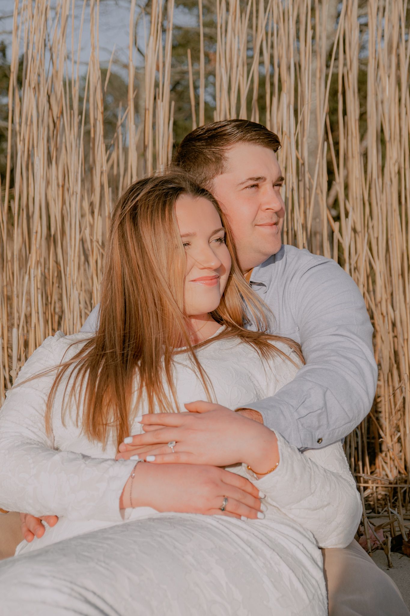 Couple cuddling together in front of tall golden beach grass during a sunset engagement session.