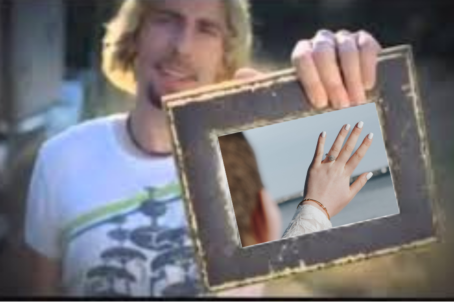 A man with long blonde hair holds a wooden picture frame displaying a hand with rings outdoors.