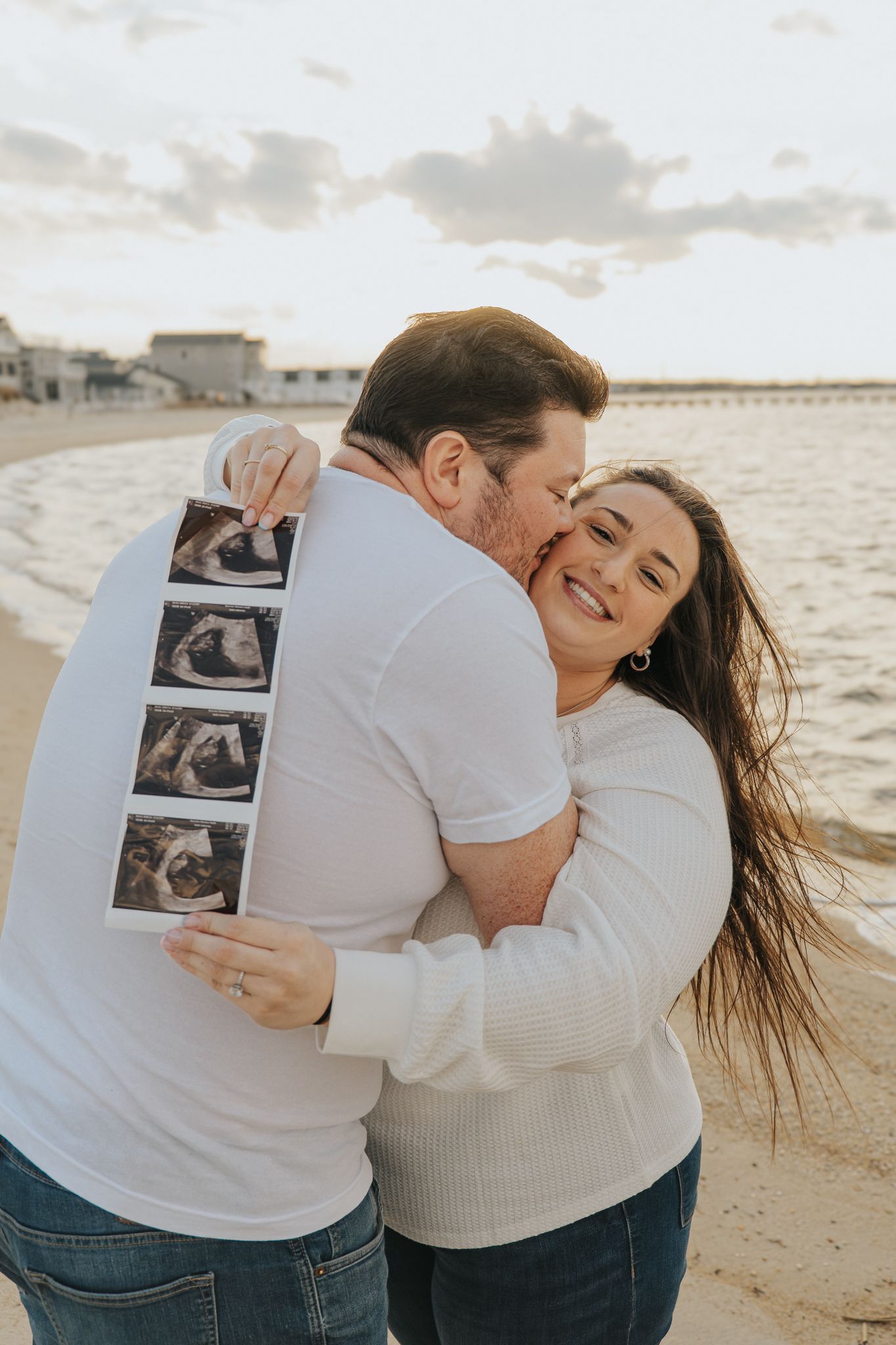 Happy couple hugging on beach holding ultrasound photos, announcing pregnancy at sunset near pier.
