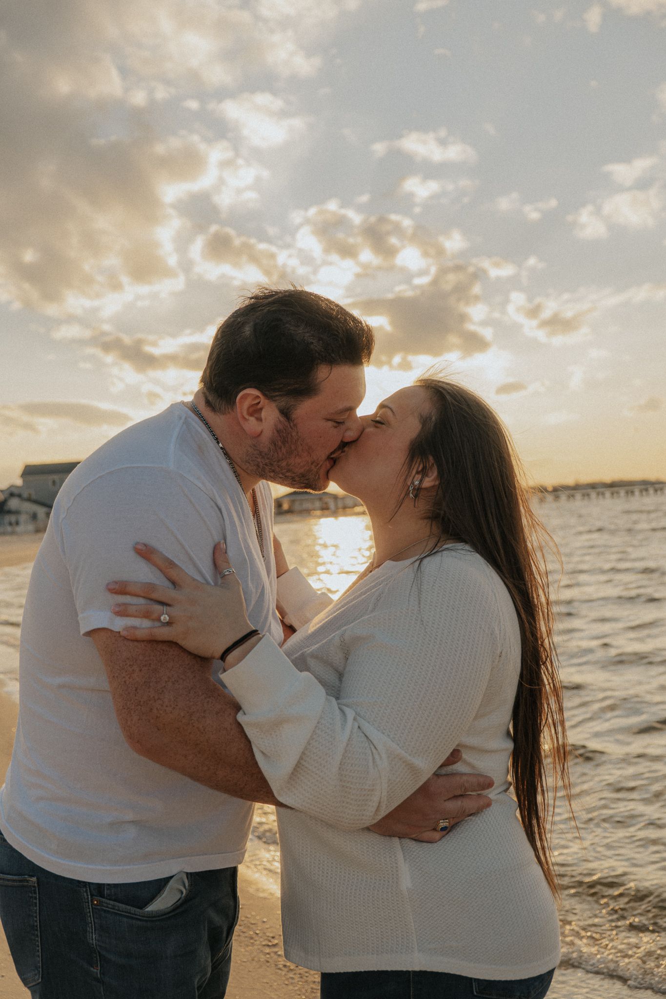 Couple kissing tenderly on a sandy beach at sunset, with a pier and warm golden sky in the background.
