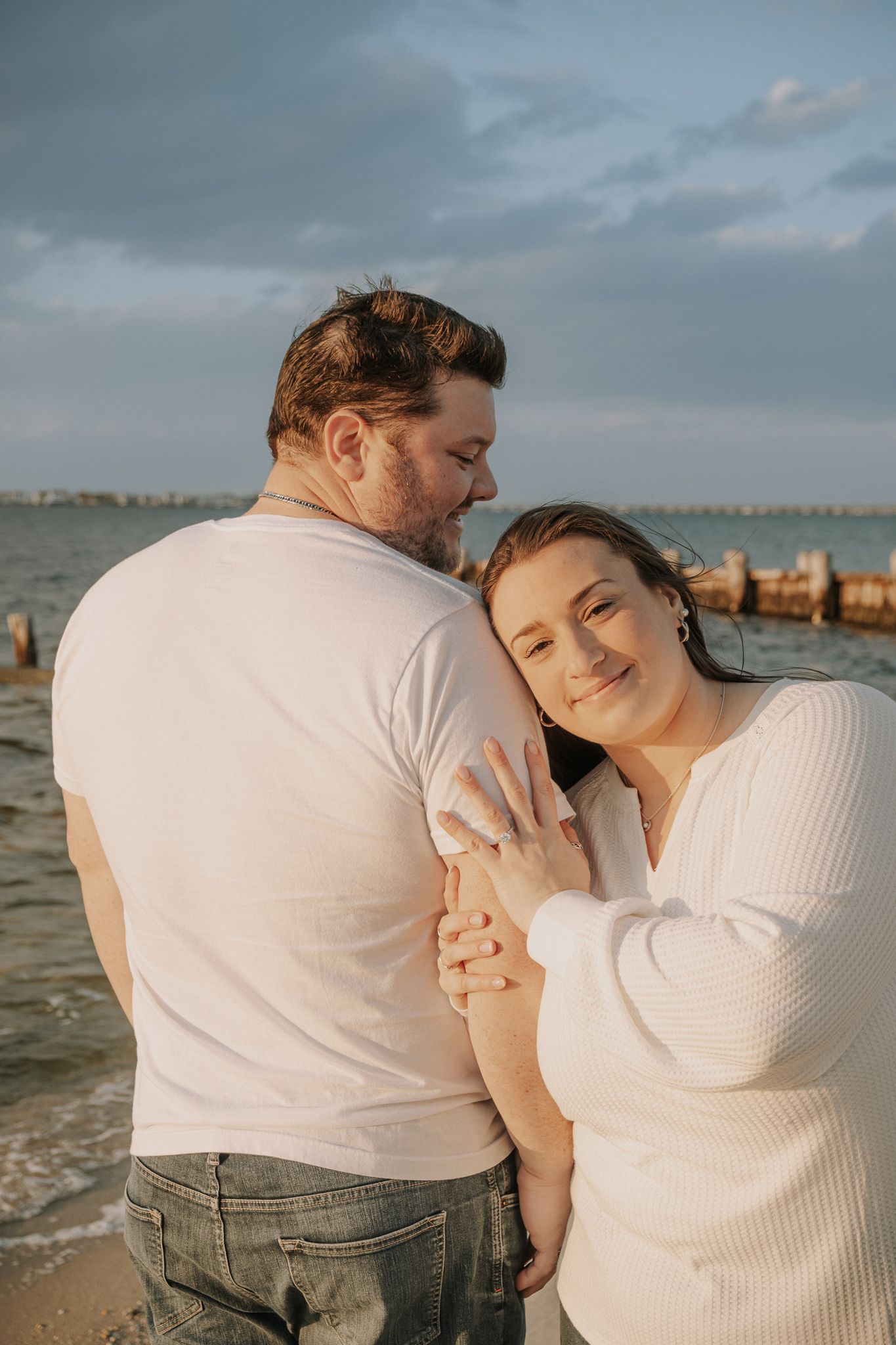 Couple embracing on a beach at golden hour, woman in white sweater rests head on mans shoulder near the ocean.