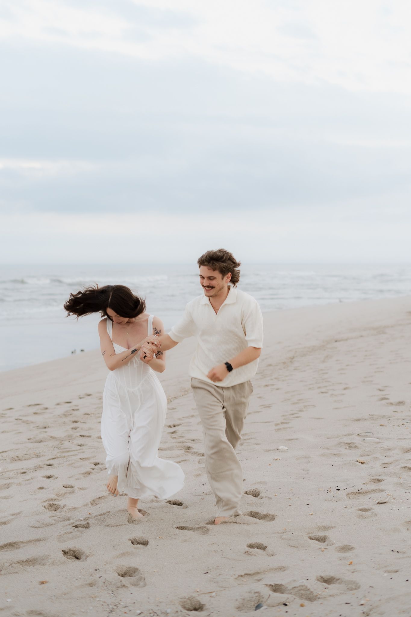 A joyful couple in white outfits runs barefoot along a sandy beach, holding hands and laughing together.
