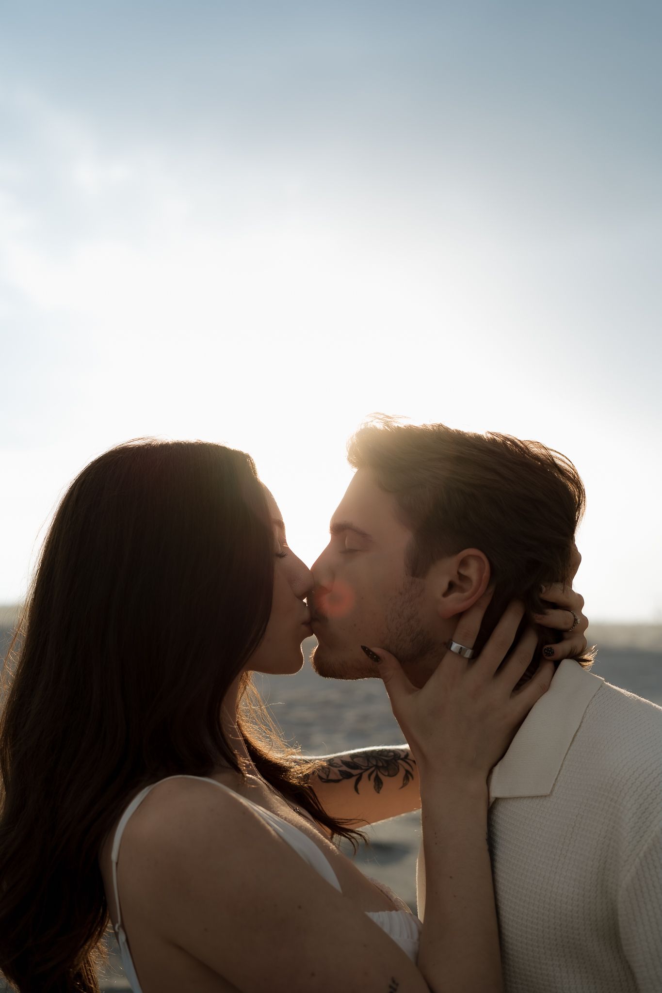 Romantic couple sharing a kiss at sunset on beach, woman with tattoo embracing mans neck in warm backlight.