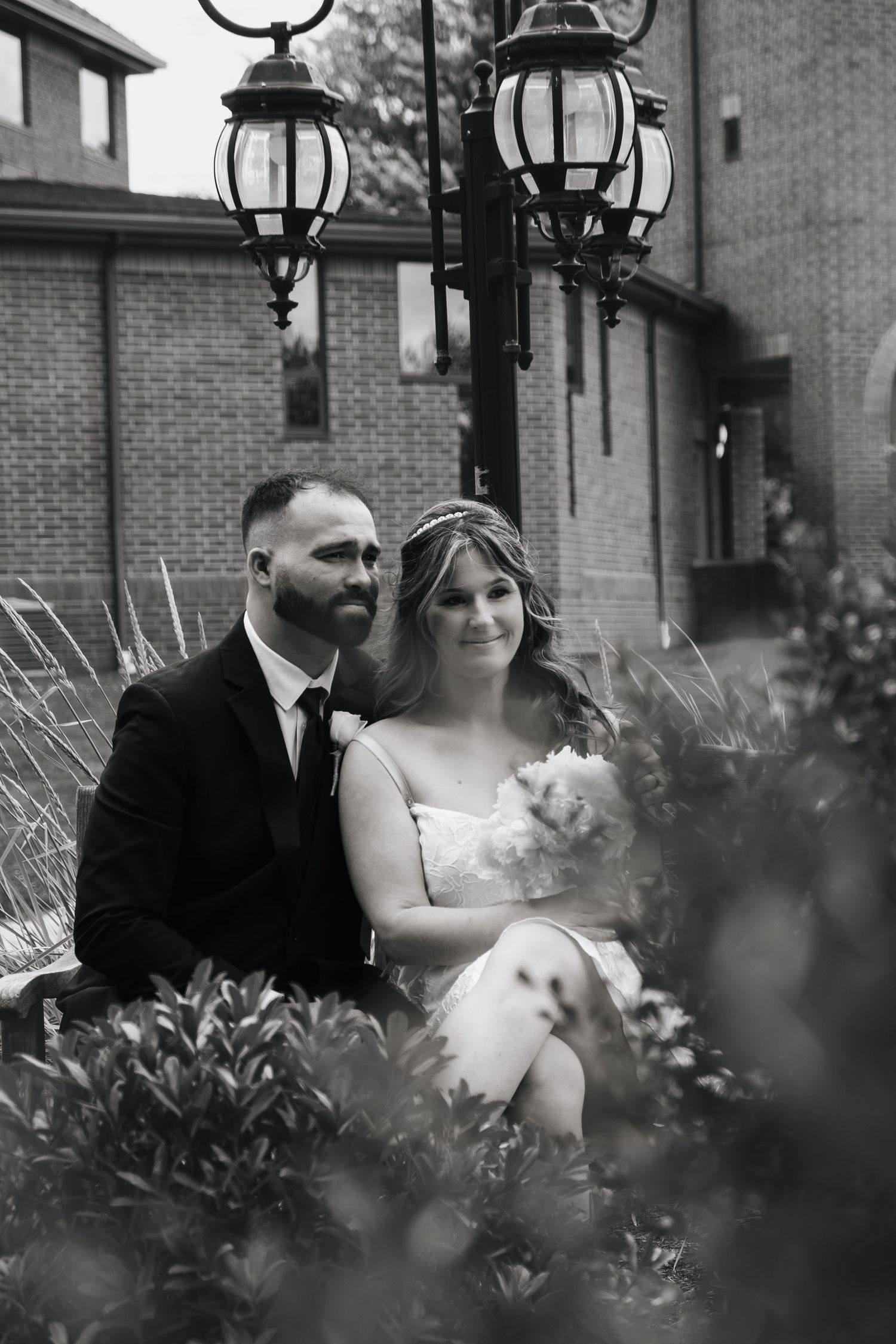 Black and white portrait of bride and groom seated together near vintage lanterns and brick building.