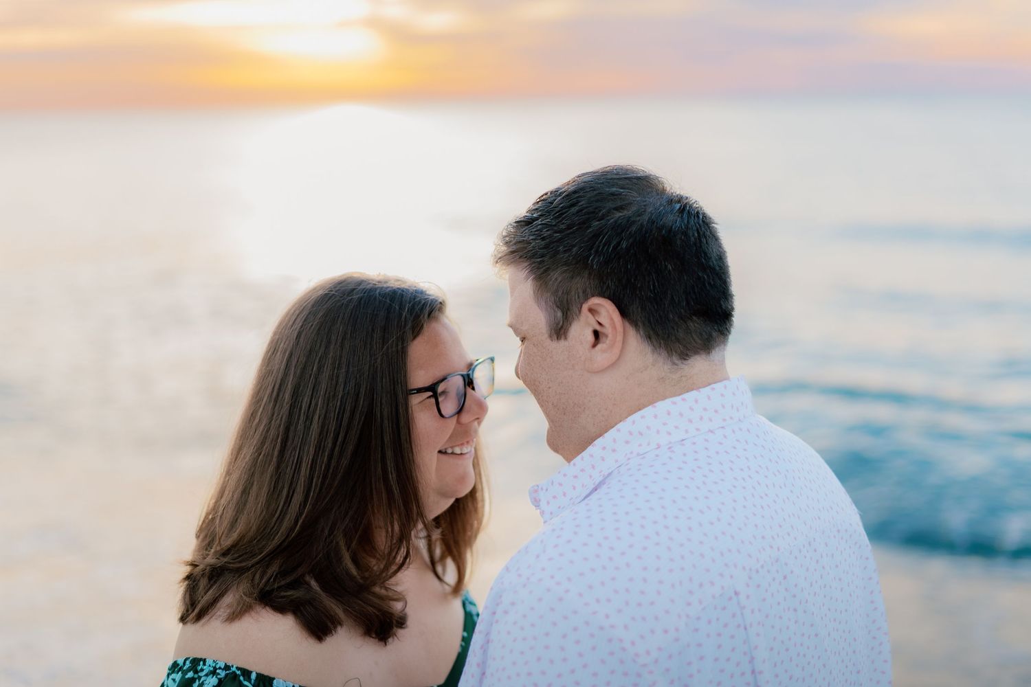Couple sharing a romantic moment at sunset on the beach, woman wearing glasses smiling at her partner.