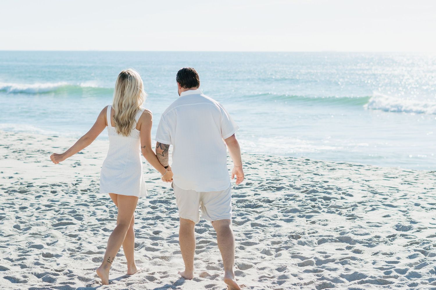 Couple holding hands walking barefoot on a sandy beach toward ocean waves, both wearing white outfits on a sunny day.