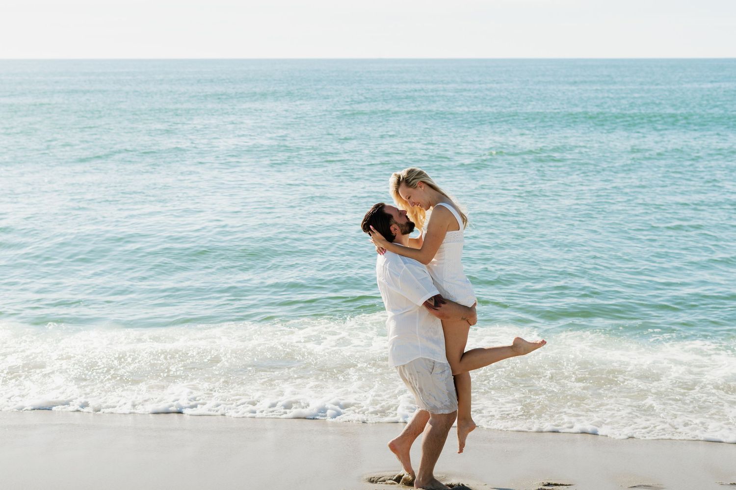 Couple embracing and kissing on a sunny beach with ocean waves, wearing white outfits in a romantic moment.