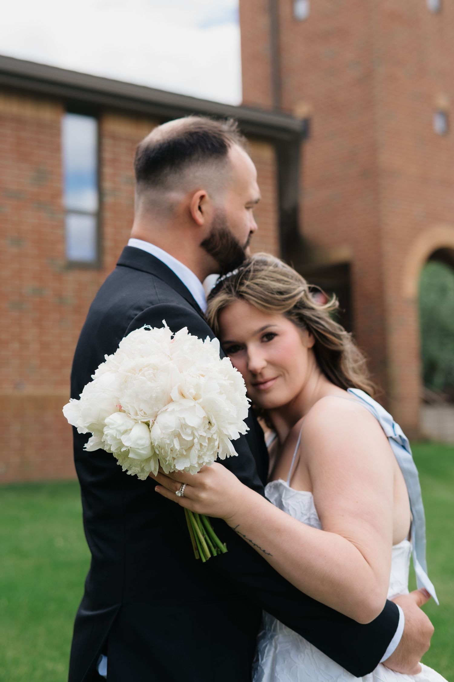 Bride holding white peony bouquet leans against groom in black suit outside brick church on wedding day.
