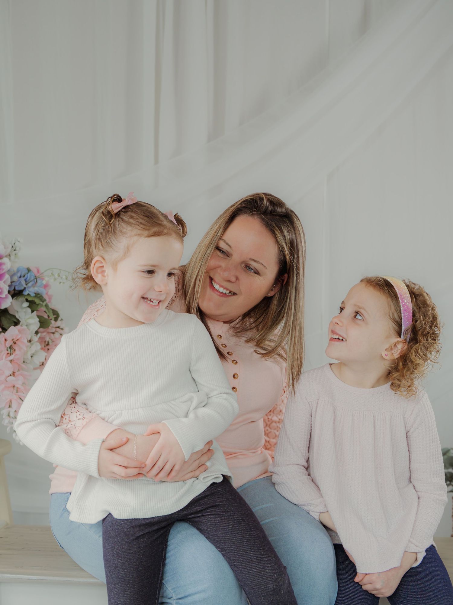 A smiling mother poses with her two young daughters in pink outfits against a bright white background.