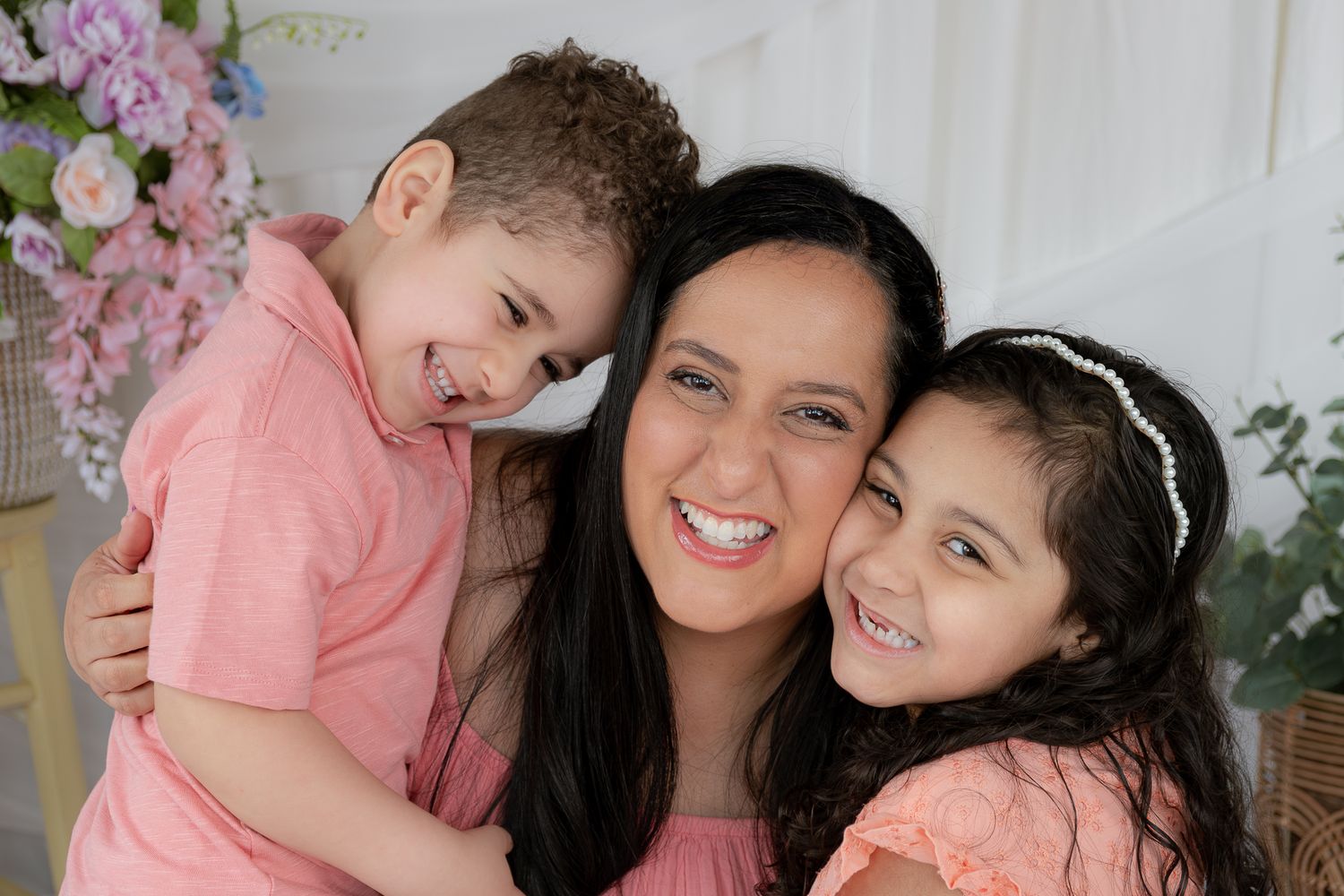 A dark-haired mother smiles warmly while hugging her young son and daughter in pink outfits.