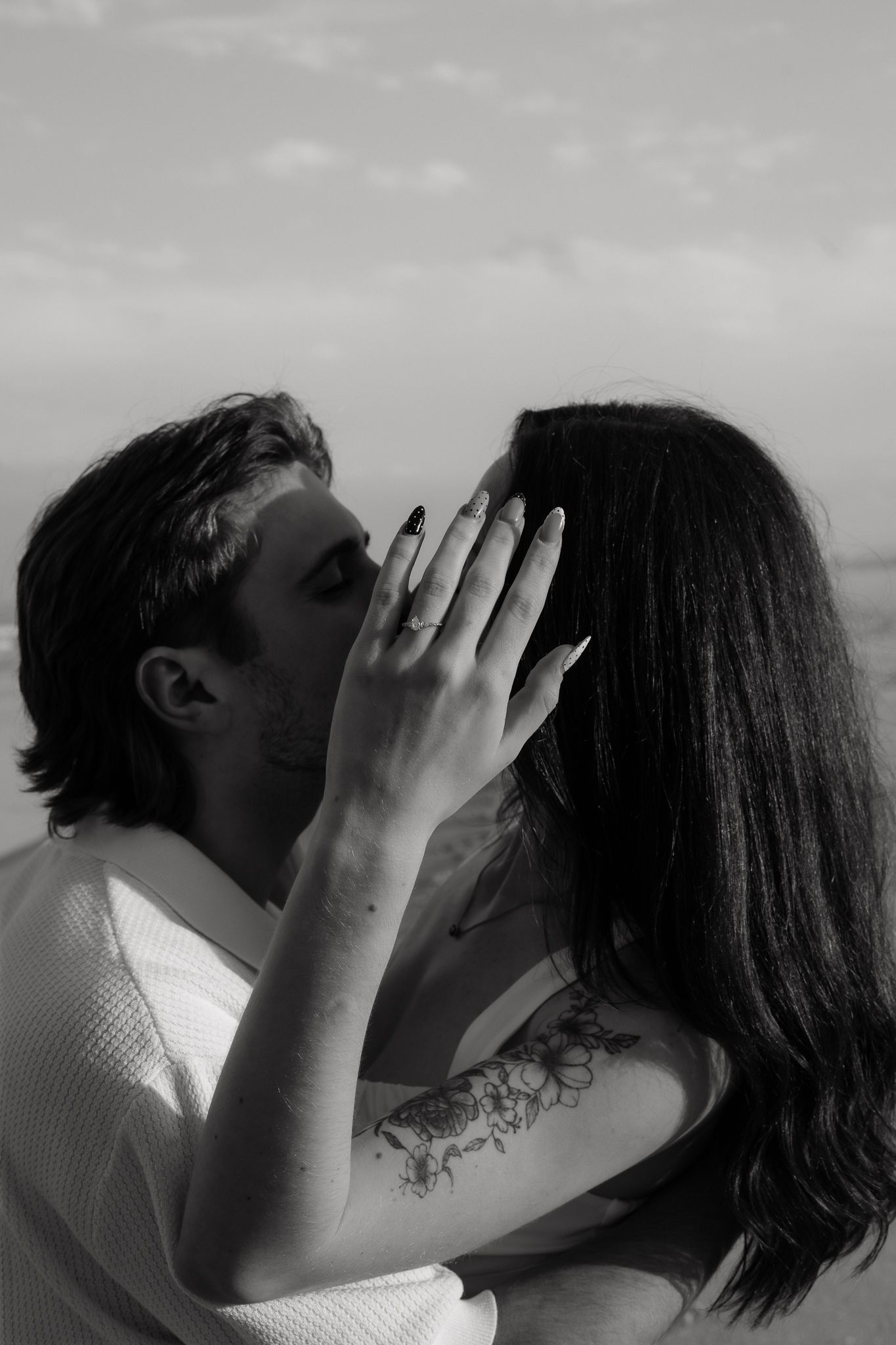 Black and white photo of a couple sharing a tender kiss outdoors, woman with floral tattoo gently holds mans face.