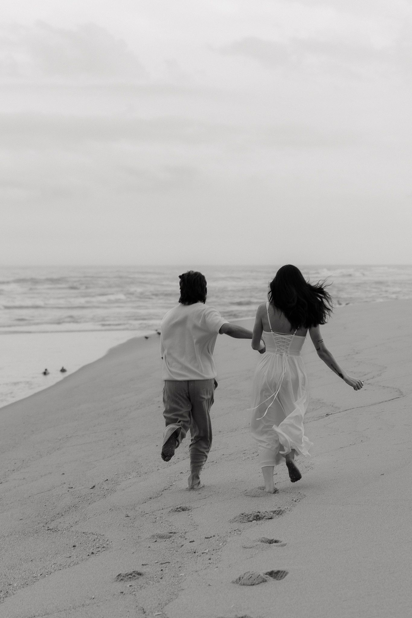 Black and white photo of a couple holding hands running along a sandy beach toward ocean waves.