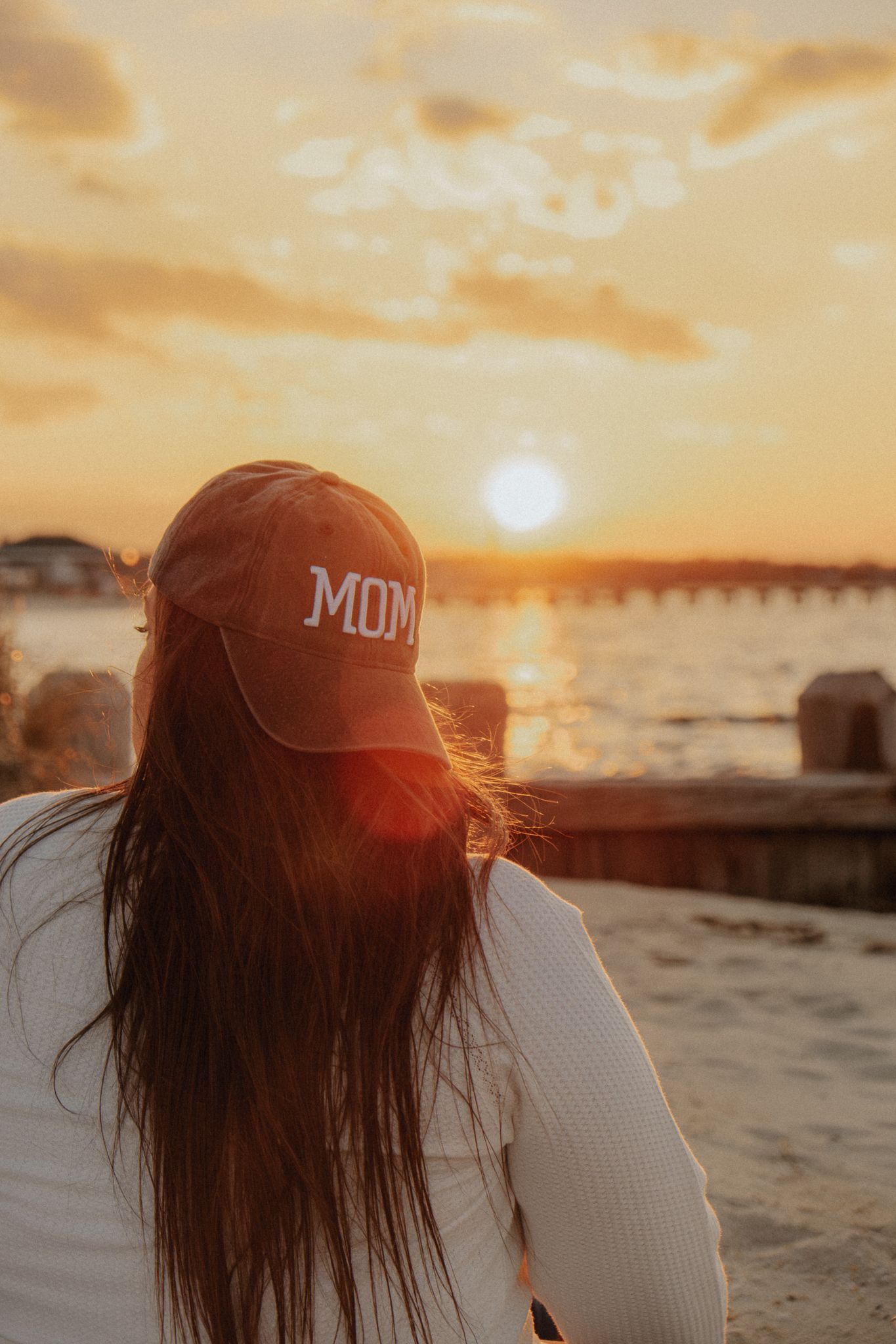Woman wearing a MOM hat watching a golden sunset over the ocean from a sandy beach.