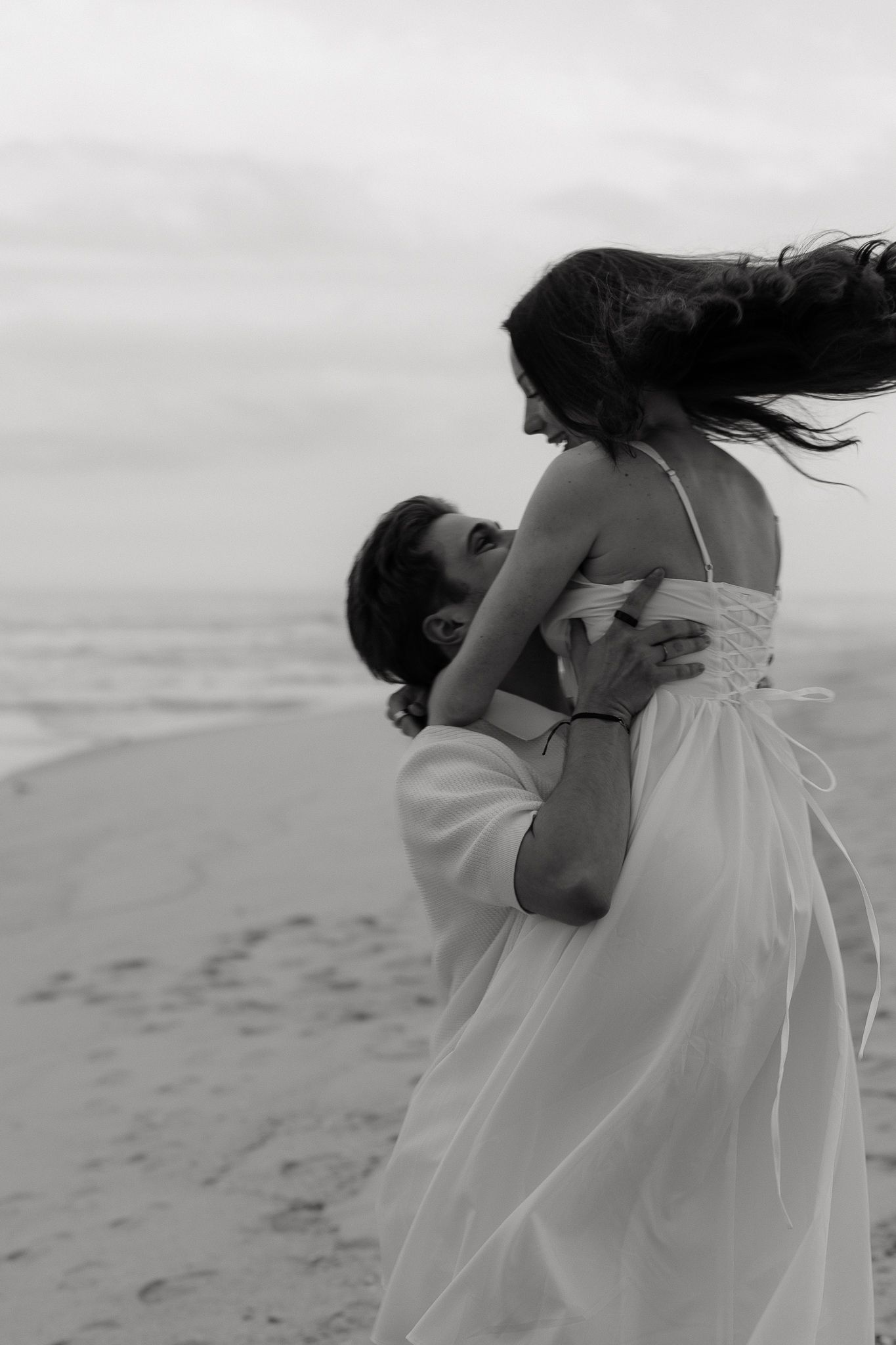 Black and white photo of a couple embracing on a beach, woman in white dress with hair blowing in the wind.