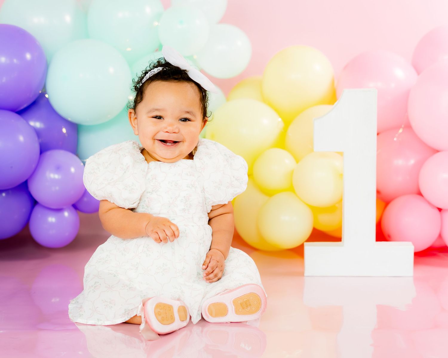 Baby girl in white dress smiles at her first birthday photoshoot with pastel rainbow balloons and number one.