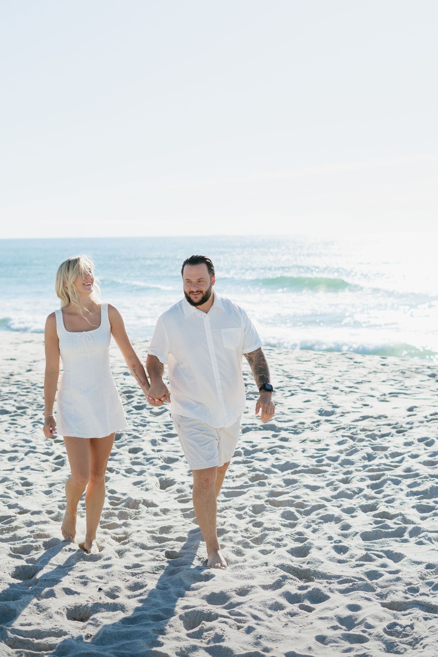 Couple in white outfits holding hands and walking on a sandy beach with ocean waves in the background.