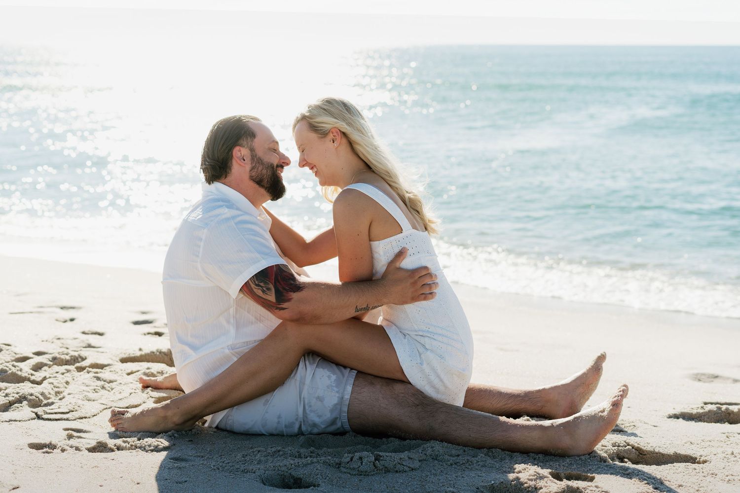 Couple sitting together on sandy beach, sharing a romantic moment with ocean waves in the background.