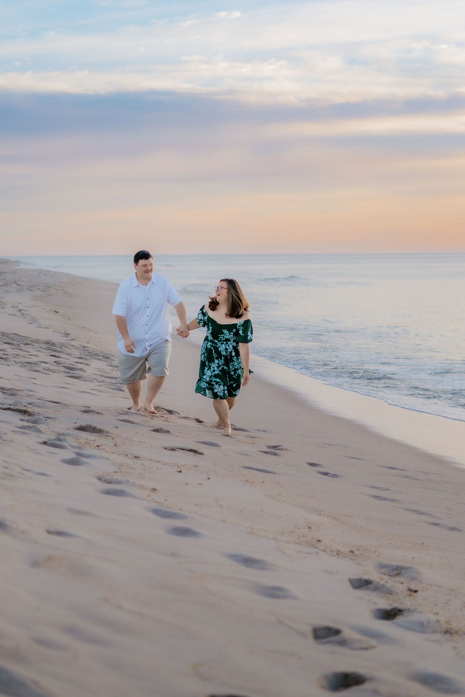 Couple holding hands walking barefoot on sandy beach at sunset, man in white shirt and woman in green floral dress.