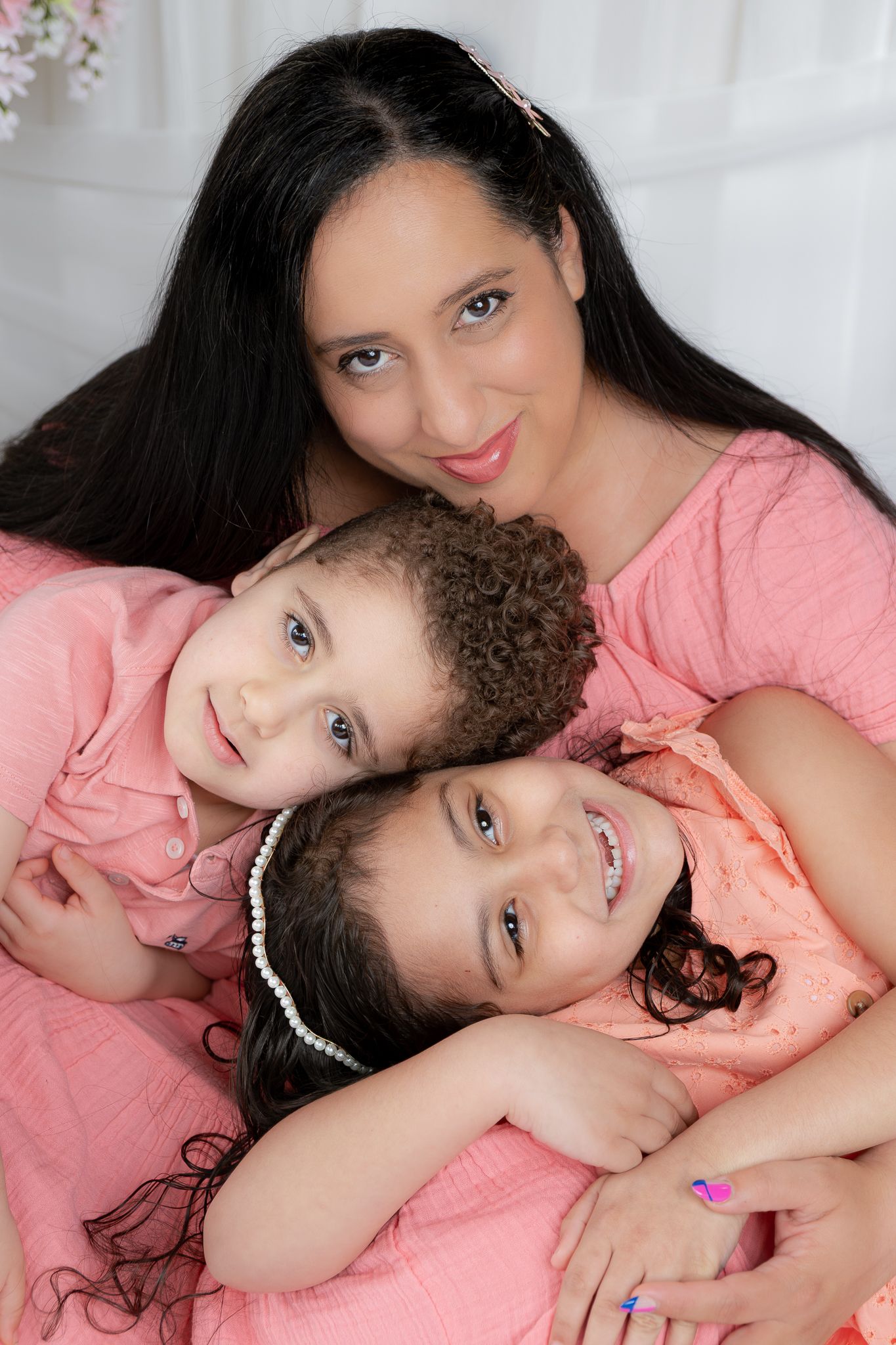 A mother with long dark hair poses with her two young daughters, all wearing matching coral pink outfits.