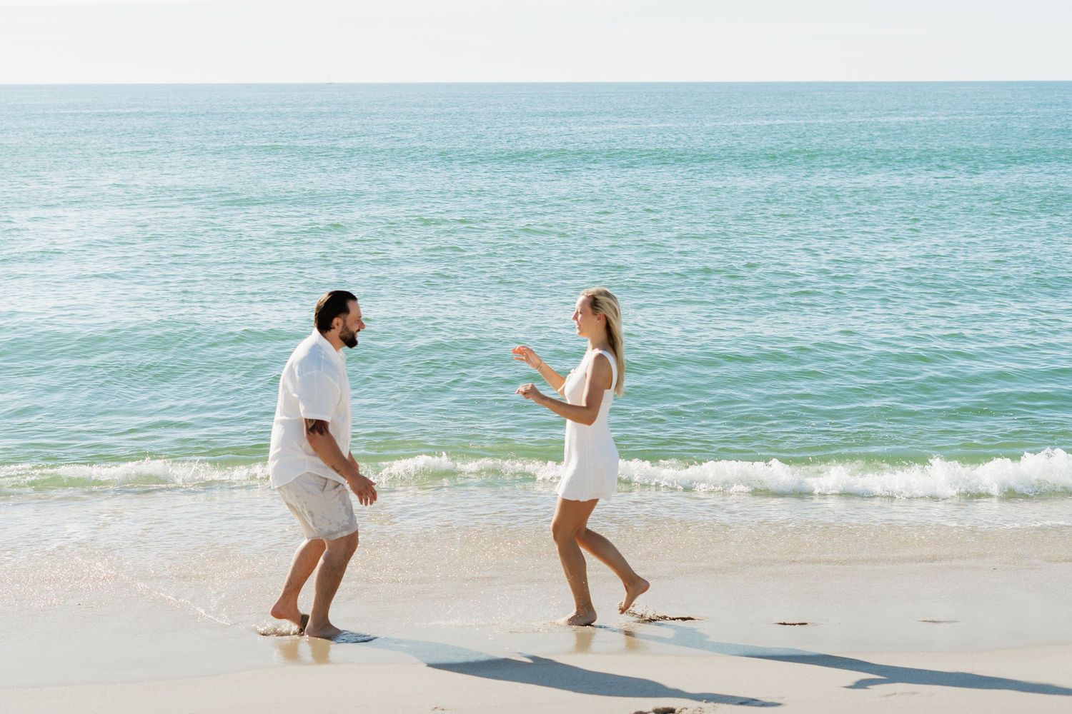 Couple in white outfits playing on a sunny beach with turquoise ocean waves in the background.
