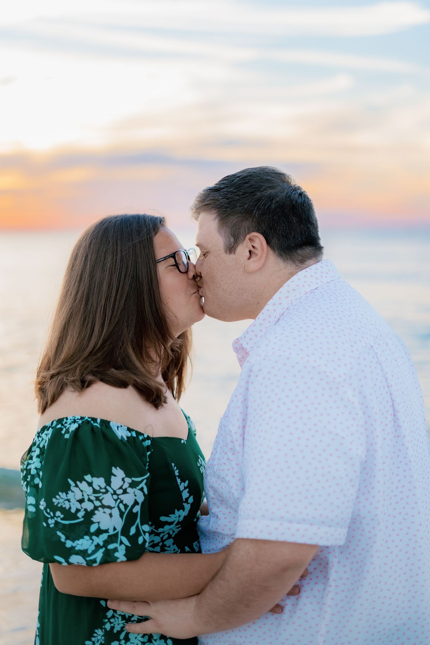 Couple sharing a romantic kiss on the beach at sunset, woman in green floral dress and man in white shirt.