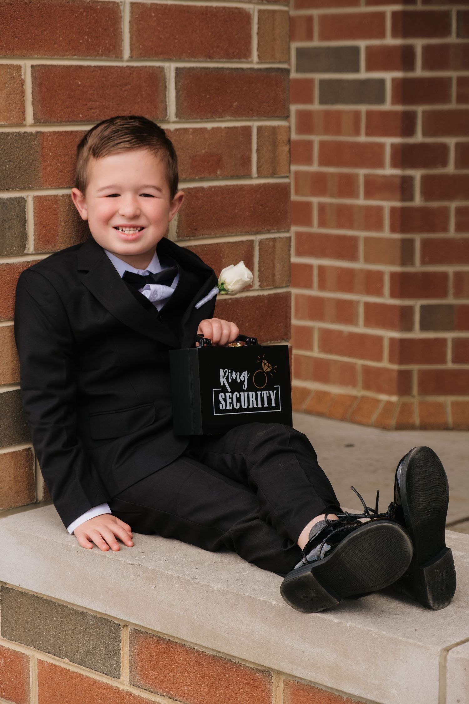 Young ring bearer in black suit sitting against brick wall holding a Ring Security case at a wedding.
