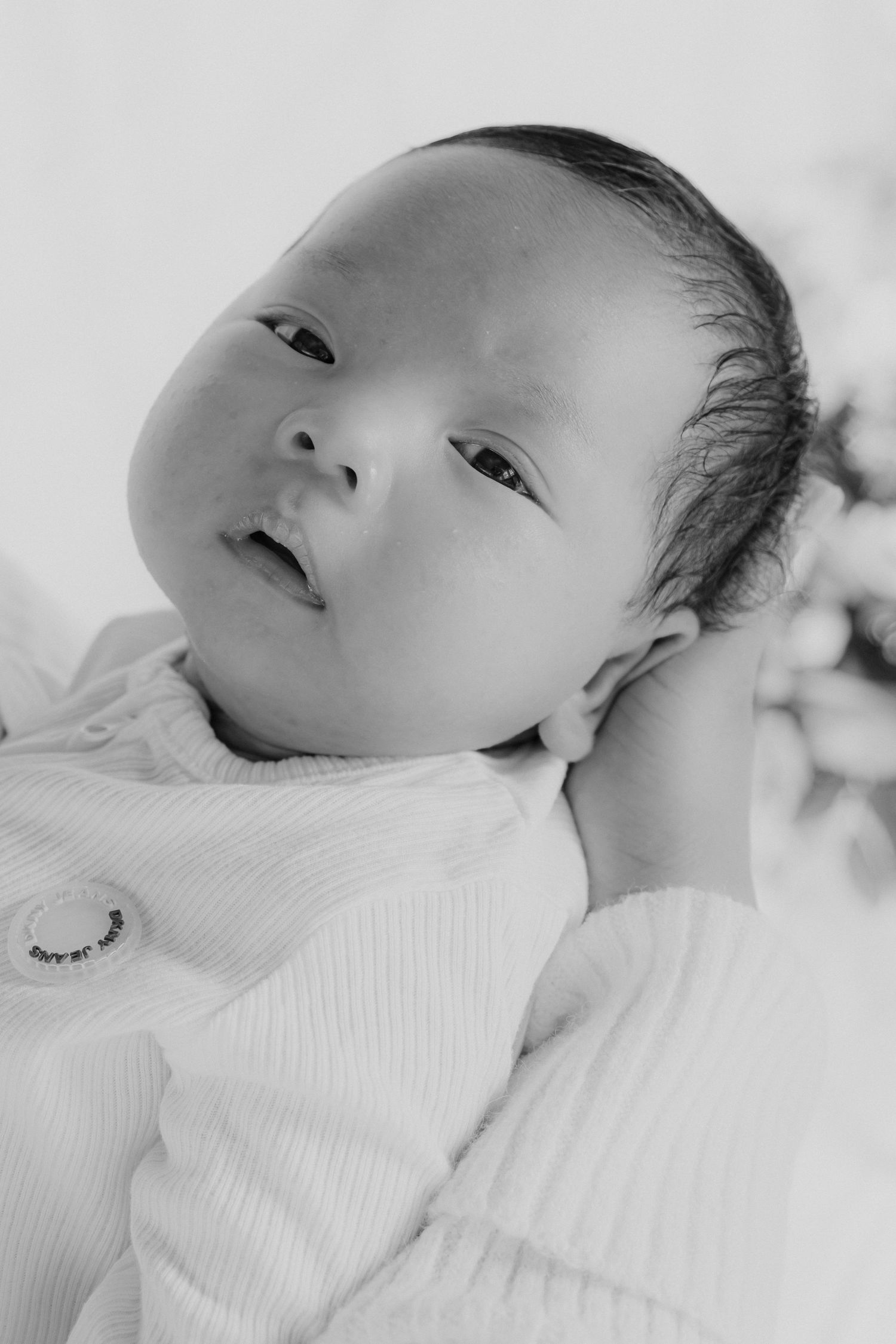 Black and white portrait of a newborn baby with dark hair, looking up with wide eyes, wearing a white ribbed outfit.
